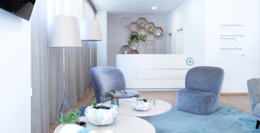 Salle d’attente lumineuse et accueillante de la clinique de fertilité Ferticentro à Coimbra avec fauteuils gris, tables basses et comptoir de réception blanc.